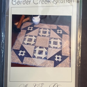May include: A quilt pattern named "Border Creek Station" with a geometric design in blue and beige. The pattern includes the text "And Sew On..." and the dimensions 61cm x 10cm.
