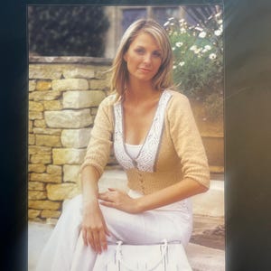 Peut inclure: Un livret de modèles Jaeger Handknits intitulé The Aqua & Trinity Collection JB39. La couverture présente un gilet en tricot beige à manches trois-quarts et boutons, avec une bordure en dentelle blanche le long du décolleté en V, porté sur un haut blanc.