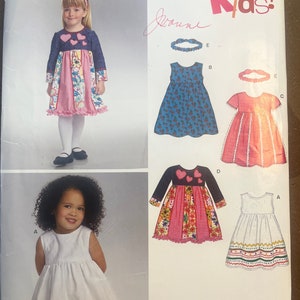 May include: A sewing pattern for children's dresses. The pattern includes five different dress styles, including a white dress with a colourful trim, a pink dress with hearts, a blue dress with flowers, a pink dress with a checked pattern, and a white dress with a colourful border. The pattern is from New Look and is size A 1/2-4.