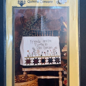 May include: A sewing machine cover with a floral design and the text "Friends are the Flowers in Life's Garden." The cover features a patchwork border with star patterns. The packaging reads "The Button Bush Quilting Company."