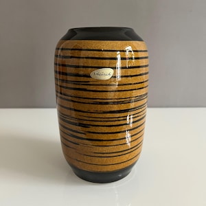 Puede incluir: Un jarrón de cerámica rayado en marrón y negro con un borde y una base negros. El jarrón tiene una etiqueta que dice "Scheurich".
