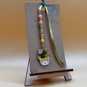 May include: A gold bookmark with a pink and green cactus charm and a beaded chain. The bookmark is on a brown card stand.