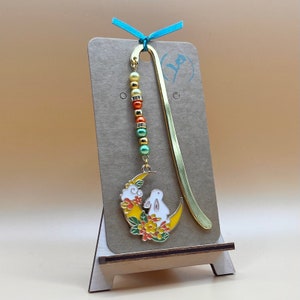 May include: A gold bookmark with a crescent moon charm featuring a white rabbit and orange and green flowers. The bookmark has a beaded chain with green, orange, and yellow beads.