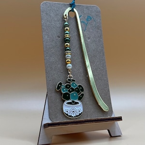 May include: A gold bookmark with a green and white succulent charm. The charm is attached to a chain of gold and green beads with white pearls.