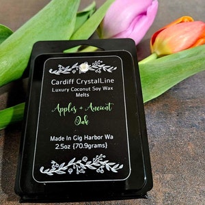 May include: A black wax melt package with the text "Cardiff CrystalLine" and "Apples + Ancient Oak." The package contains 2.5oz (70.9 grams) of luxury coconut soy wax melts. The image also features green, pink, and orange tulips.