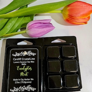 May include: Black wax melts in a plastic container, labeled "Eucalyptus Mint" with text. A purple and orange tulip and green leaves are in the background. The label says "Cardiff CrystalLine Luxury Coconut Soy Wax Melts".