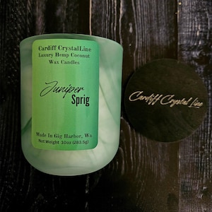 May include: A light green candle in a glass jar with a black lid. The candle has a label that reads "Juniper Sprig" and "Cardiff Crystal Line." The net weight is 10oz (283.5g).