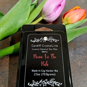 May include: A black wax melt package with the text "Picnic In The Park" and "Cardiff CrystalLine" is displayed. The package is surrounded by green leaves and pink and orange tulips. The package contains 2.5oz (70.9 grams) of wax melts.