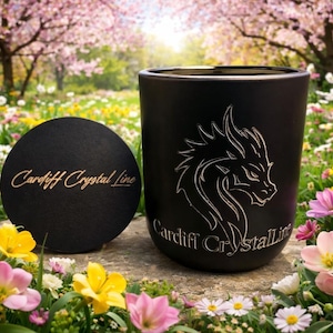 May include: Black cylindrical candle holder with a dragon design and the text "Cardiff Crystal Line". A matching black coaster with the same text is next to the candle holder. The items are set on a stone surface surrounded by flowers.