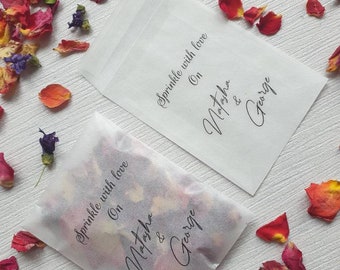 Può includere: Due bustine di carta bianca piene di petali di fiori colorati, sparsi su una superficie bianca. Le bustine sono stampate con le parole "Sprinkle with love On Natasha & George" in elegante corsivo.