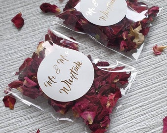 Può includere: Due sacchetti di plastica trasparenti pieni di petali di rosa rossa. Ogni sacchetto ha un adesivo circolare bianco con lettere dorate che recitano "Mr & Mrs Whittouck".