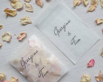Può includere: Due sacchetti di carta bianca con i nomi Georgina & Tim scritti su di essi. I sacchetti sono pieni di petali di rosa.