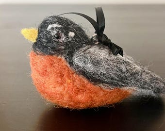 May include: A handcrafted felt bird ornament with a black ribbon for hanging. The bird features a vibrant orange breast, gray and black wings, and a yellow beak. This decorative item is suitable for seasonal decor.