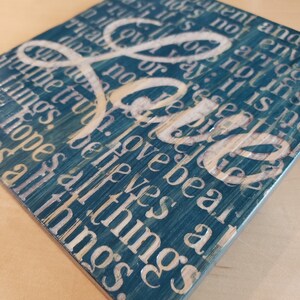 May include: A teal blue wooden sign with white lettering that reads "Love bears all things, believes all things, hopes all things, endures all things".