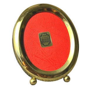 May include: A small, oval-shaped, gold-colored brass picture frame with a red backing. The frame has a shiny finish and rests on two small, round feet. A sticker on the frame reads "hand polished SOLID BRASS".