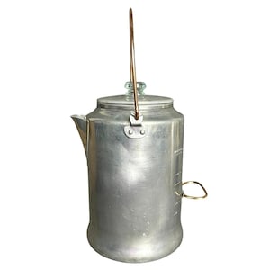 May include: A vintage aluminum coffee pot with a copper-colored handle and a glass knob on the lid. The pot has a spout and a side handle. It has measurement markings on the side.