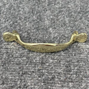 May include: A gold-colored, curved drawer pull with a textured surface. The handle has a slight arch and rounded ends, designed for easy gripping. This hardware is suitable for cabinets, drawers, or other furniture.