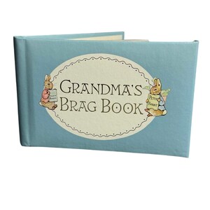 May include: A light blue photo album with a central cream-colored oval label featuring the text Grandma's Brag Book. The cover is decorated with two illustrations of rabbits, one on each side of the oval, each holding a small rectangular sign.