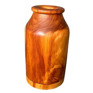 May include: A handcrafted wooden vase with a natural, warm brown color. The vase has a wide base, a cylindrical body, and a flared top. The wood grain is visible, adding to its rustic charm. This decorative vase is suitable for home decor.