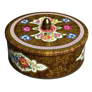 May include: A round metal tin with a wood grain pattern, featuring a central brass-colored knob on the lid. The surface is decorated with colorful floral motifs in red, pink, blue, and yellow, accented by white panels and a pink geometric border.