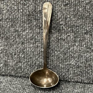 May include: A silver ladle with a long, slender handle and a rounded bowl. The ladle is sitting on a gray, textured surface.