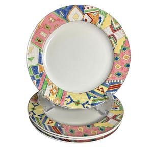 May include: A stack of white ceramic plates with a colorful geometric pattern around the rims. The pattern includes pink, blue, yellow, and green shapes. The plates are stacked on a clear plastic stand.