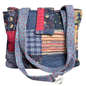 May include: A quilted patchwork tote bag featuring a variety of fabric patterns including blue plaid, red stripes, and floral motifs. The bag has two shoulder straps and a central flap closure with a heart-shaped metal tag labeled Donna Sharp.