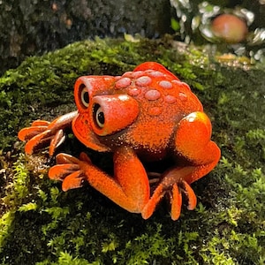 Op de afbeelding: Een feloranje kikkerbeeldje met grote, prominente ogen en een textuurrijke, hobbelige rug. De kikker bevindt zich in een gehurkte houding met de ledematen onder het lichaam gevouwen, wat een levendige, verzadigde kleur en afwerking toont.