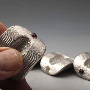 Pode incluir: Close-up de vários anéis prateados com uma superfície texturizada e uma pequena pedra preciosa vermelha. Os anéis têm uma forma orgânica e abstrata. Um anel é segurado em uma mão, outros ao fundo.