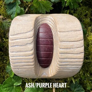 Puede incluir: Un objeto cilíndrico de madera con un exterior de madera de fresno clara y una ranura vertical prominente que contiene una pieza lisa de madera de corazón púrpura. El texto ASH/PURPLE HEART está impreso en blanco en la parte inferior.
