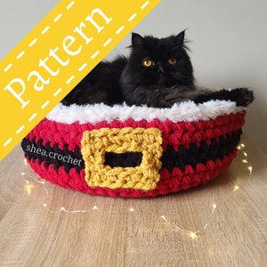 May include: A red, black, and white crocheted cat bed with a yellow buckle detail. The bed is shaped like a Santa Claus belt and has a white fluffy lining. A black cat is sitting inside the bed.