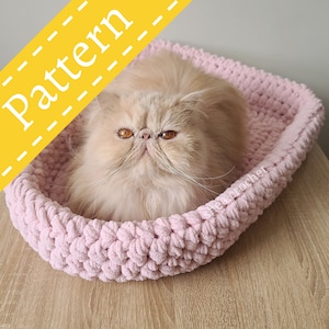 Puede incluir: Una cama para gatos de crochet rosa con un gato esponjoso de color marrón claro descansando dentro. La cama está hecha con un hilo grueso y texturizado y tiene una forma redondeada.