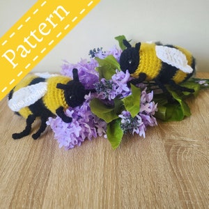 May include: Two crocheted bumblebees with black and yellow stripes and white wings are nestled in a bouquet of purple lilac flowers. The bees are handmade and have a cute, whimsical look.