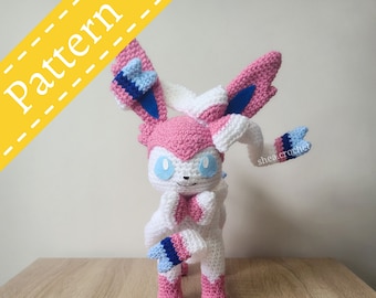 May include: A crocheted pink and white Pokemon Sylveon plush toy with blue and light blue accents. The toy has a curled tail and is standing on a wooden surface.