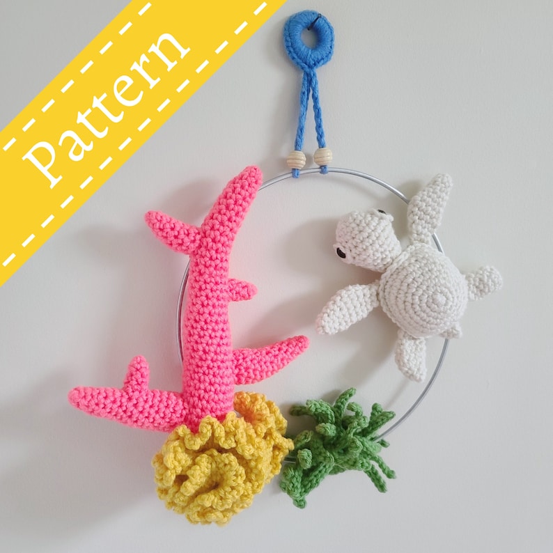 May include: A crocheted sea turtle, coral, and seaweed hanging mobile. The mobile is made of white, pink, yellow, and green yarn and is attached to a silver metal ring.