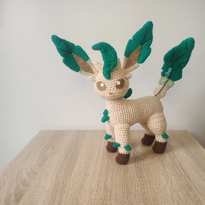 May include: A crocheted plush toy of a light brown Eevee with green leaf-like appendages. The toy has brown eyes and a light brown body with green accents.