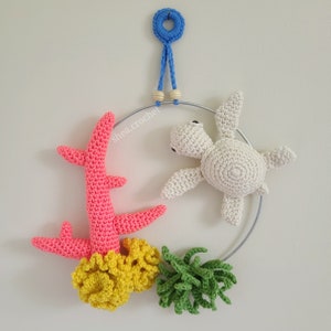 May include: A crocheted sea turtle, coral, and seaweed mobile hanging from a metal ring. The mobile is made of white, pink, yellow, and green yarn.