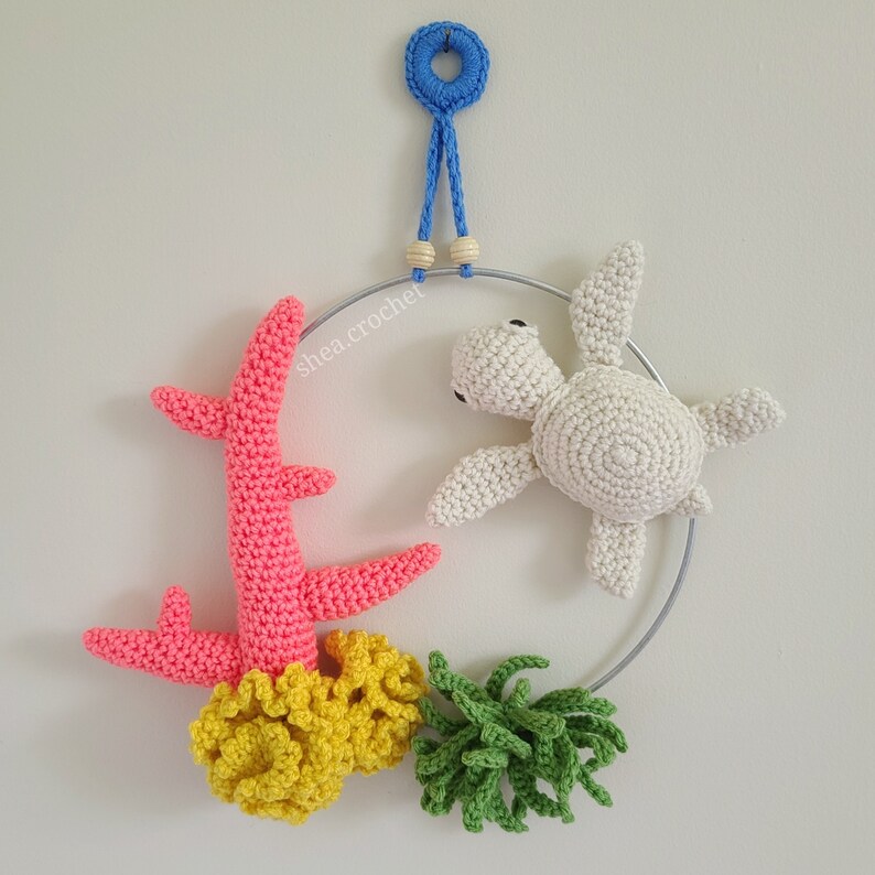 May include: A crocheted sea turtle, coral, and seaweed mobile hanging from a metal ring. The mobile is made of white, pink, yellow, and green yarn.