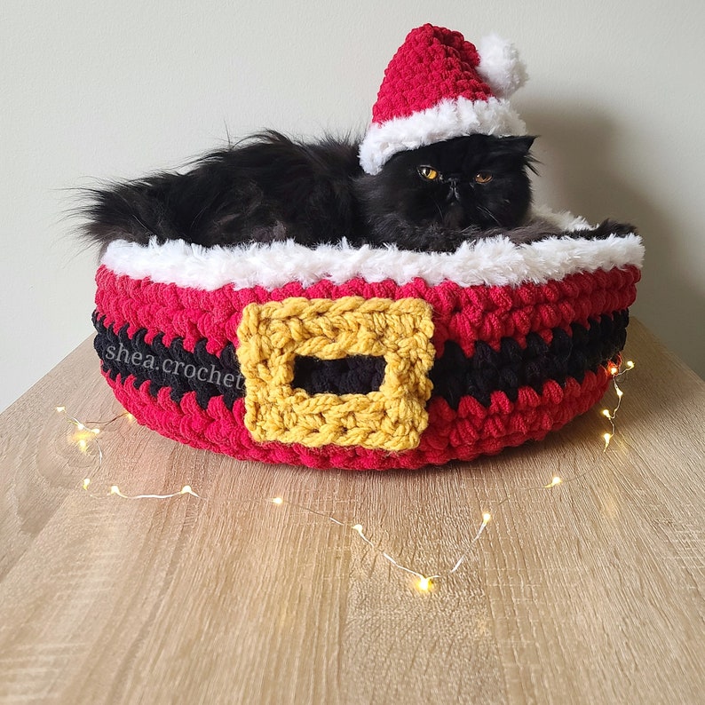 Puede incluir: Un gato negro con un gorro de Pap&aacute; Noel de crochet rojo y blanco est&aacute; acurrucado en una cama de gato de crochet rojo, blanco y negro con un cintur&oacute;n de crochet amarillo. La cama est&aacute; sobre una superficie de madera con luces de hadas blancas.