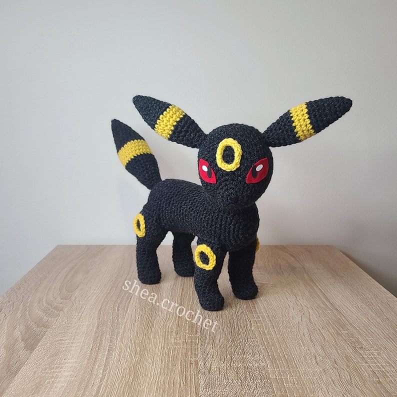 K&ouml;nnte beinhalten: Eine schwarze und gelbe geh&auml;kelte Pokemon-Figur, Umbreon, mit roten Augen. Die Figur steht auf einer Holzoberfl&auml;che.