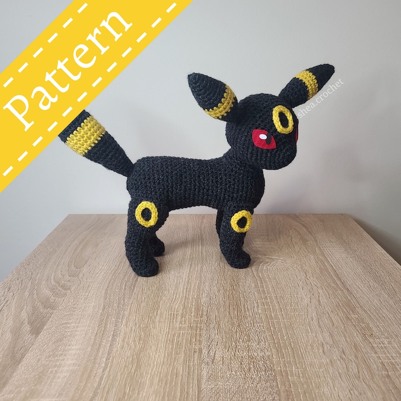 K&ouml;nnte beinhalten: Schwarzes H&auml;kel-Amigurumi eines Pok&eacute;mon-Charakters, Umbreon. Der Charakter hat gelbe Akzente an seinen Ohren und Beinen. Der Charakter hat rote Augen. Die geh&auml;kelte Figur steht auf einer hellbraunen Holzoberfl&auml;che. Der Text "shea.crochet" befindet sich im Hintergrund.