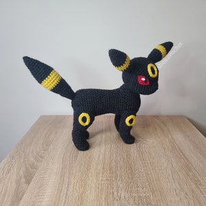 K&ouml;nnte beinhalten: Schwarzes und gelbes geh&auml;keltes Pokemon Umbreon Pl&uuml;schtier. Das Spielzeug hat rote Augen und gelbe Akzente an den Ohren und Beinen. Das Spielzeug steht auf einer Holzoberfl&auml;che. #sheacrochet