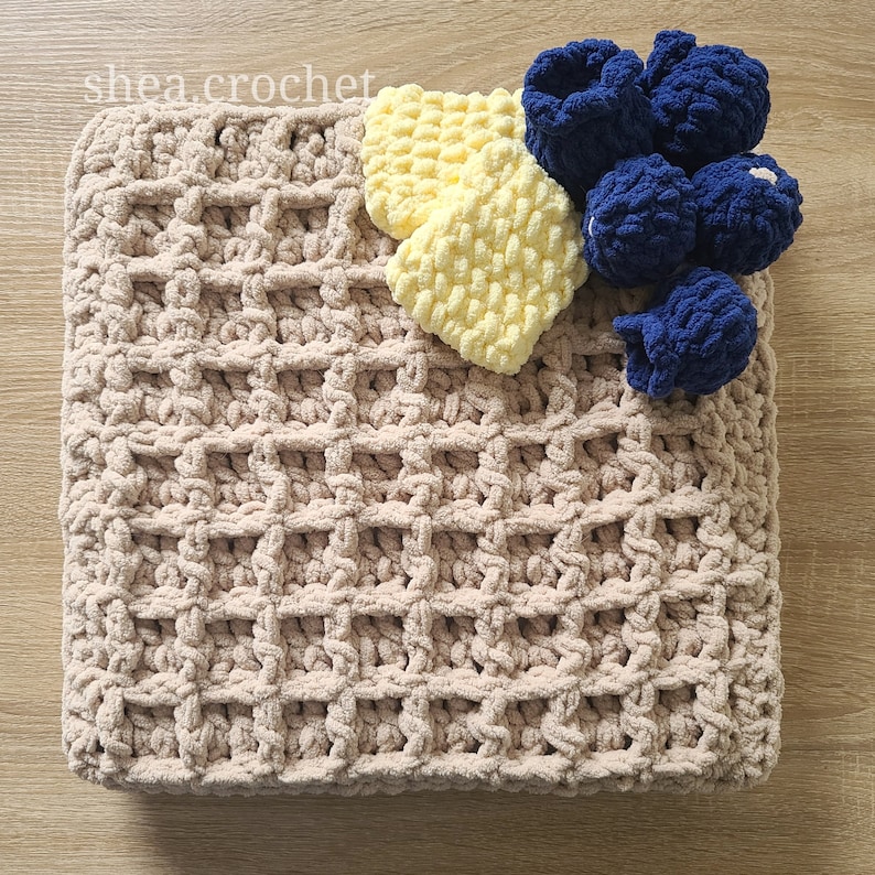 May include: A beige crocheted blanket with a waffle stitch pattern. The blanket is topped with a yellow crocheted baby bootie and a cluster of blue crocheted baby booties.