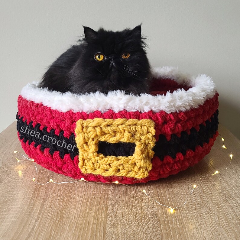 Puede incluir: Una cama para gatos de crochet roja y negra con forma de cintur&oacute;n de Pap&aacute; Noel con una hebilla de crochet amarilla. Un gato negro est&aacute; sentado dentro de la cama.