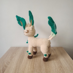 May include: A crocheted plush toy of a light brown Eevee evolution with green leafy accents. The toy is standing on a light brown wooden surface.