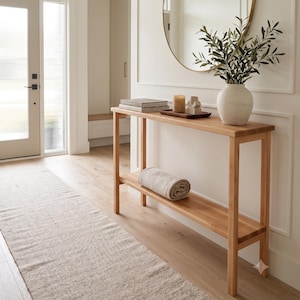 May include: A light wood console table with a lower shelf featuring a rolled beige textile, a white ceramic vase with olive branches, a dark wooden tray with a candle and a small sculpture, and a round gold-framed mirror on the wall.