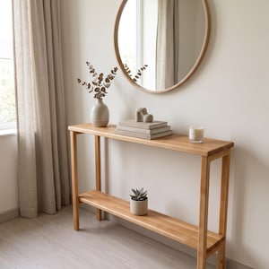 May include: A light-toned wooden console table featuring a rectangular top and a lower shelf. The table is styled with a ceramic vase containing dried branches, a stack of two books, a small abstract sculpture, a candle, and a potted succulent.