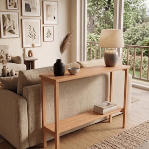 May include: A rectangular light wood console table with four straight legs and a lower shelf. The table surface holds a black vase with a dried plant stem, a small bowl, and a table lamp with a beige shade and a grey ceramic base.
