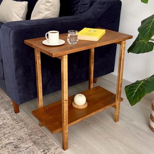 May include: A two-tiered wooden side table with a warm brown finish. The top shelf holds a white coffee cup and saucer, a glass of water, and a yellow book. The lower shelf has a candle. The table is next to a navy blue sofa.