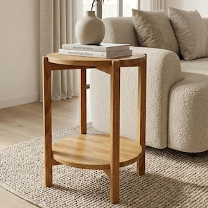 May include: A round wooden side table with a natural light brown finish, featuring a circular top and a lower shelf supported by four straight legs. Two stacked books titled Architecture Design and a beige ceramic vase are placed on the table.